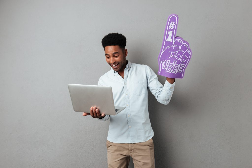 Excited young african man looking at laptop computer screen and wearing foam finger isolated over gray background